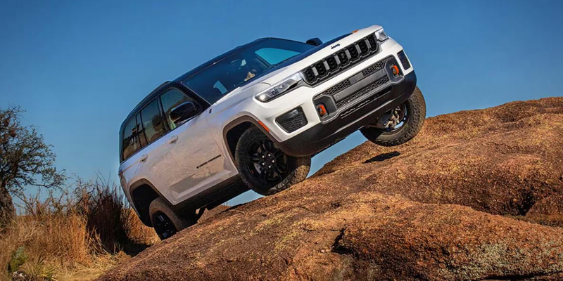 White 2026 Jeep Grand Cherokee SUV climbing over rocky terrain, showcasing off-road capability, rugged tires, and a bold front grille against a clear blue sky.