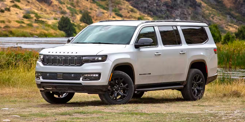 White 2026 Wagoneer SUV parked near a riverside landscape, featuring a bold front grille, black wheels, and a spacious full-size design with rolling hills in the background.